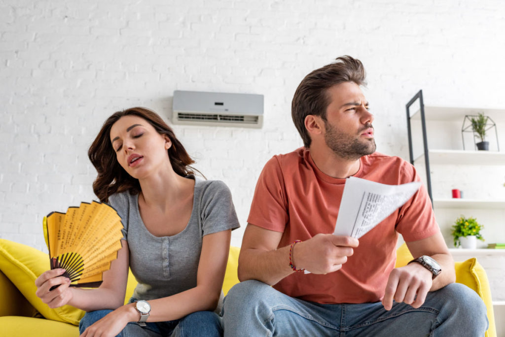 A man and woman using a fan to cool off in a home with an AC unit on the wall behind them.